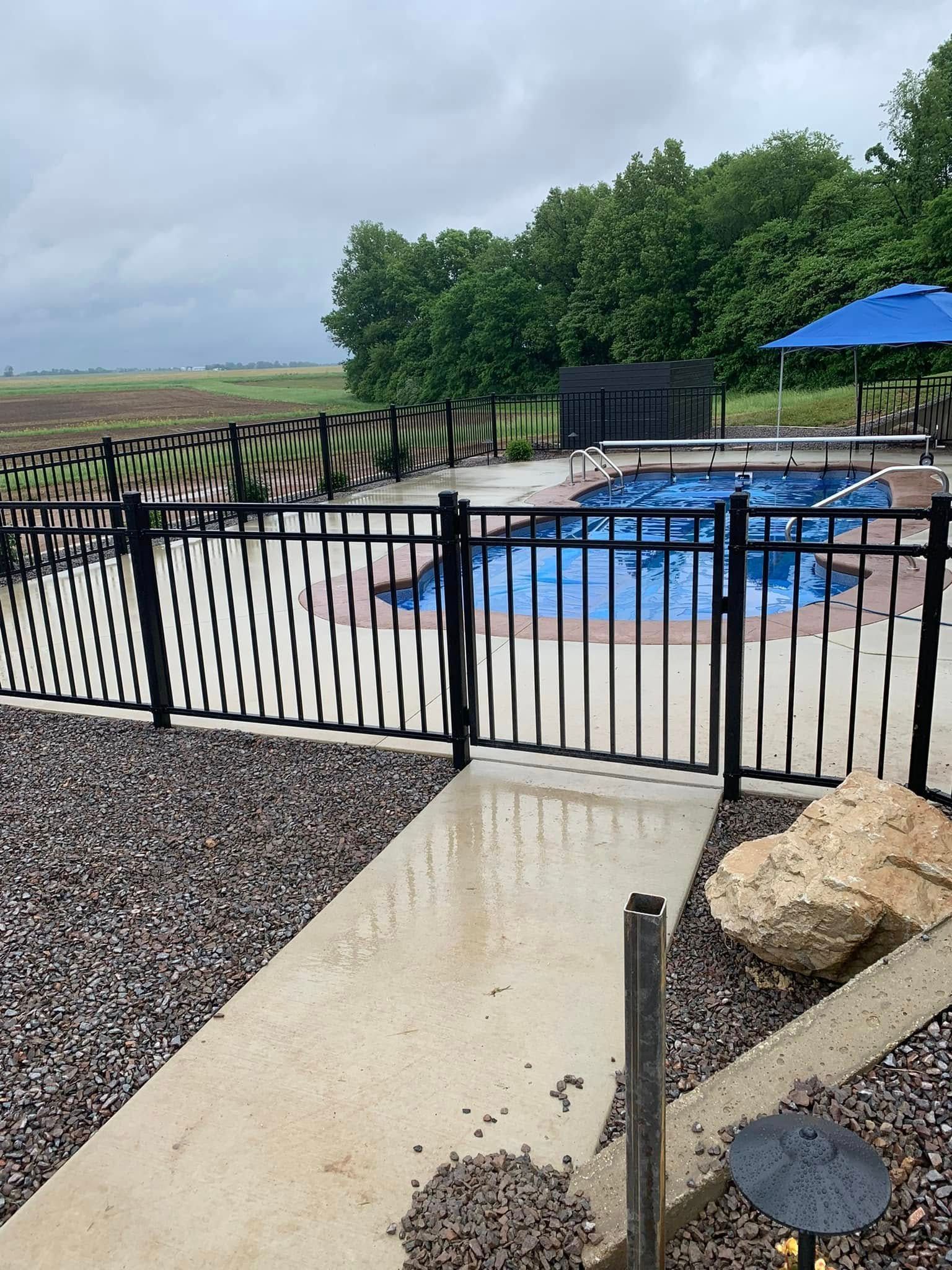 A backyard swimming pool with a blue cover, surrounded by a black metal fence and gravel landscaping on a cloudy day.