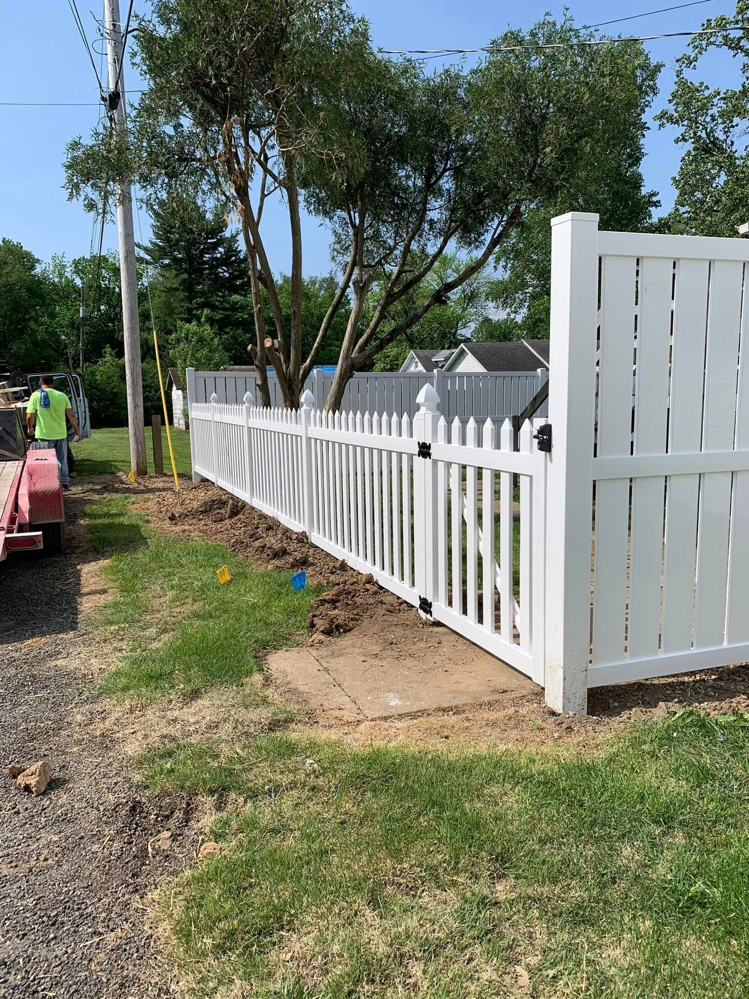 A white vinyl picket fence stands in a grassy yard, partially obscured by a tree, with a worker visible on the left.
