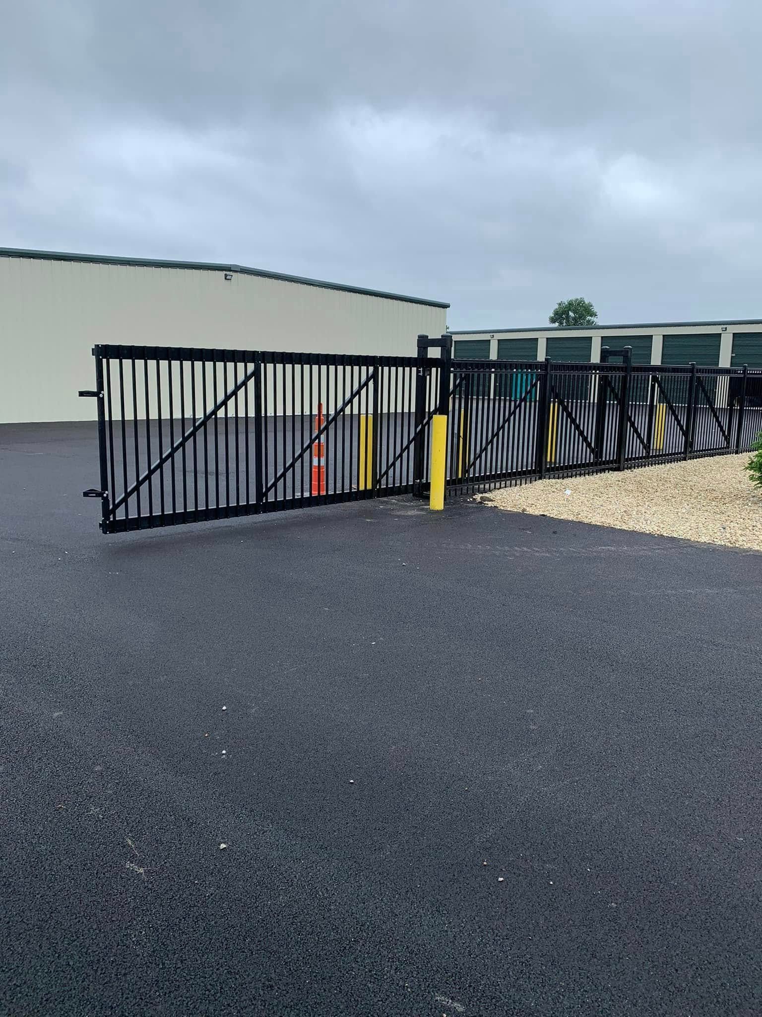 A black metal sliding gate, partially open, installed on a paved lot in front of storage units under a cloudy sky.