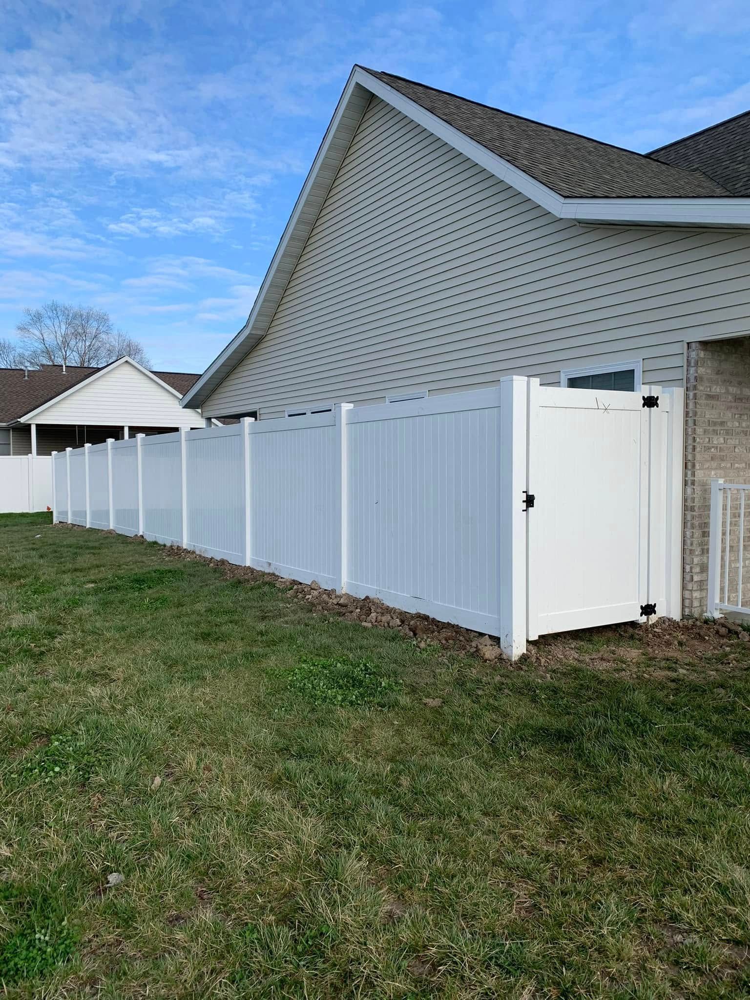 A white vinyl privacy fence extends along the side of a beige house with a light-colored shingle roof under a blue sky.