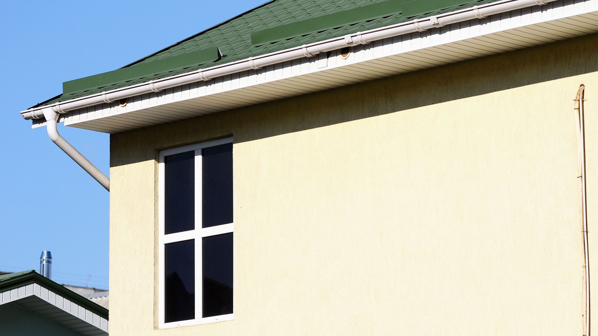 A tall, rectangular window with a white frame on a light yellow wall under a green roof overhang against a blue sky.