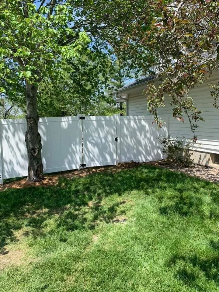 A white vinyl fence stands in a grassy backyard next to a tree and the side of a light-colored house.