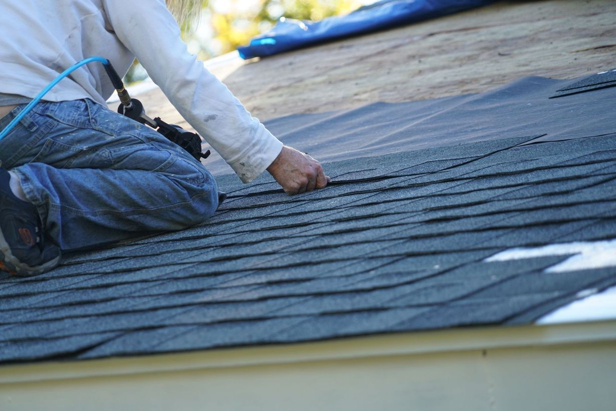 A person kneels on a sloped roof, using a nail gun to install grey asphalt shingles over a black underlayment.