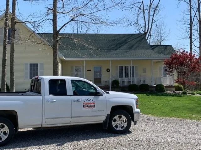 A white pickup truck with a business logo parked on a gravel driveway in front of a yellow house with a front porch.