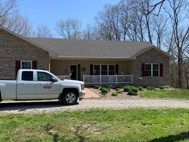 A white service truck is parked on a gravel driveway in front of a tan brick ranch-style home with red shutters.