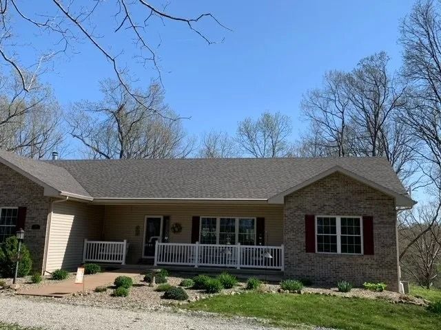 A beige brick ranch-style house with dark shingles, red shutters, and a white front porch railing under a clear blue sky.