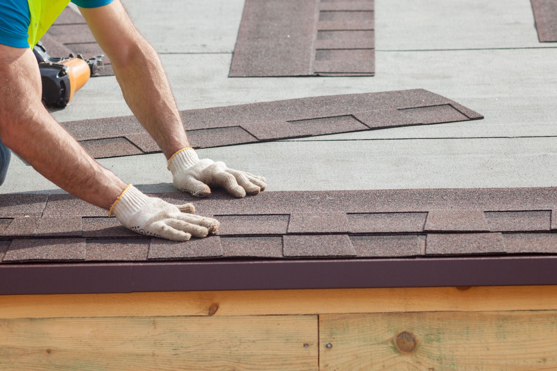 A person in work gloves installing brown asphalt shingles on a wooden roof deck.