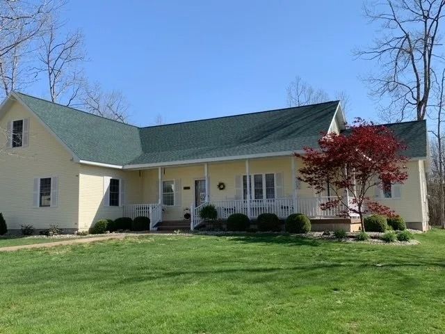 A yellow single-story farmhouse with a green roof, a front porch, and a small red tree in the front yard.