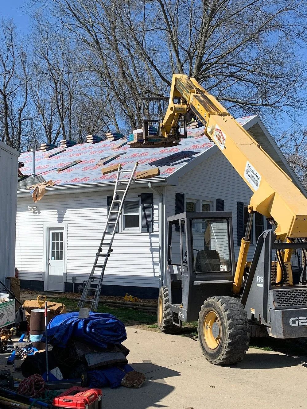 A yellow telehandler lifts roofing materials toward a house with a partially shingled roof under a clear blue sky.