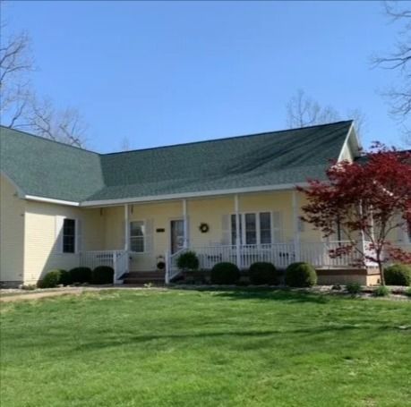 A yellow ranch-style house with a dark green roof and front porch, set against a clear blue sky and a grassy lawn.