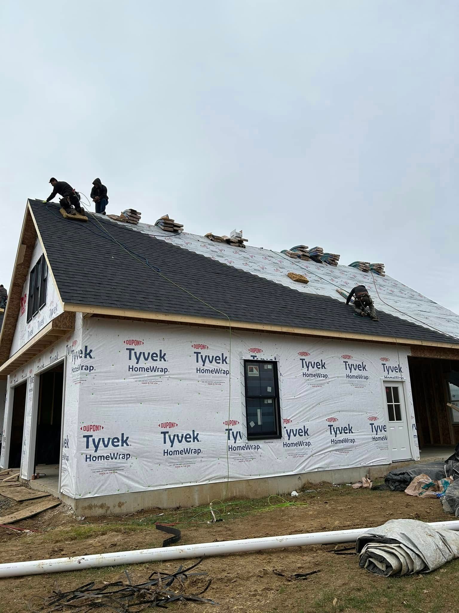 Construction workers install dark-colored shingles on the roof of a new building wrapped in Tyvek house wrap.