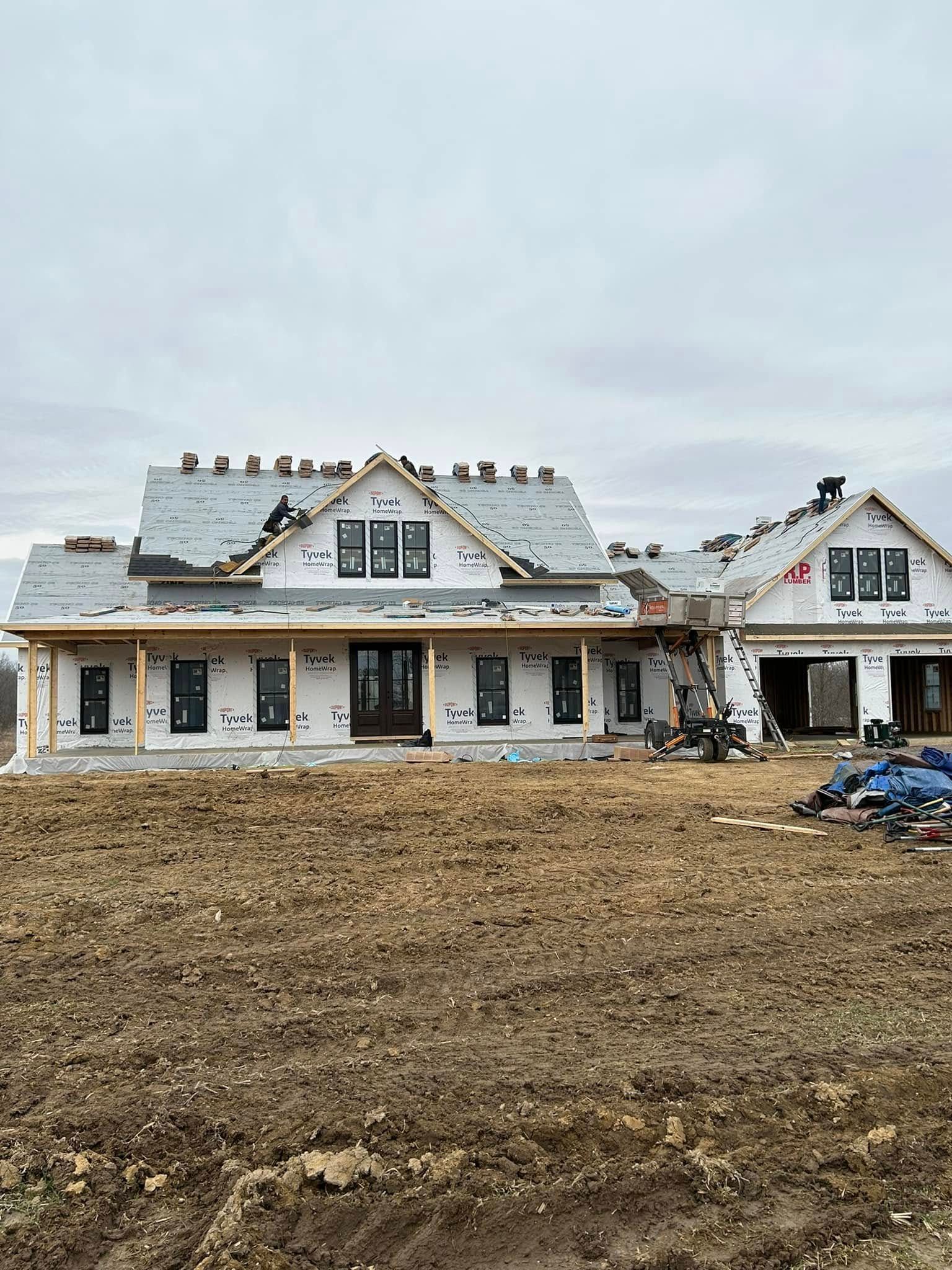 A single-story house under construction with white exterior sheathing, a gray roof, and dark window frames on a dirt lot.