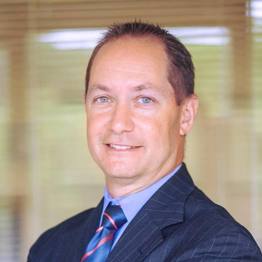 Man in suit and tie smiling, inside with window in background.