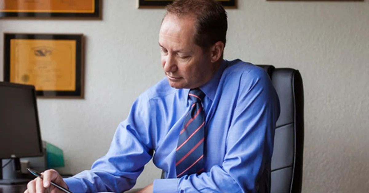 Man in blue shirt and tie, sitting at desk, writing. Office setting.