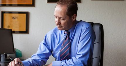 Man in blue shirt and tie, sitting at desk, writing. Office setting.