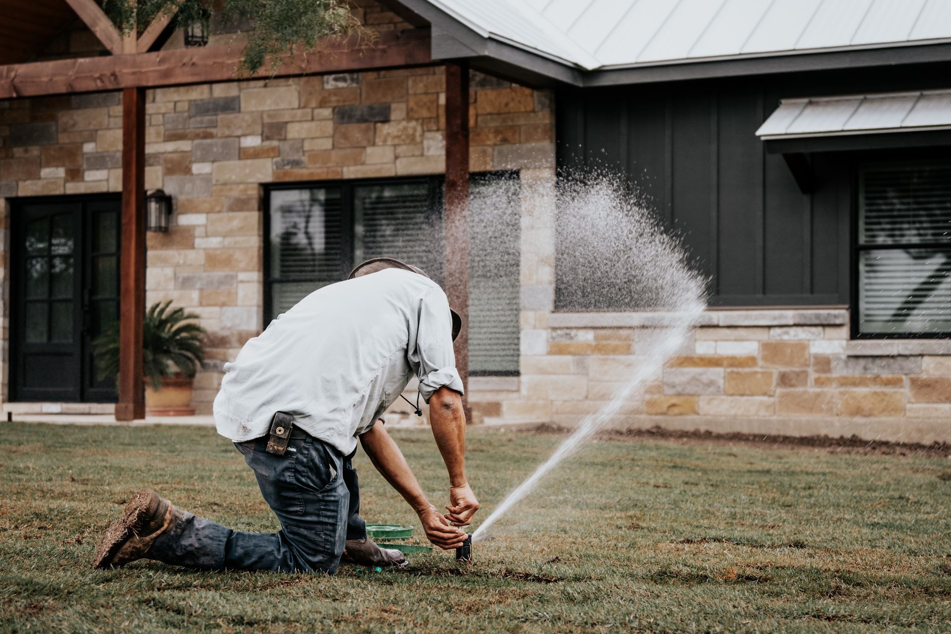 A man is kneeling down in front of a house spraying a lawn sprinkler.