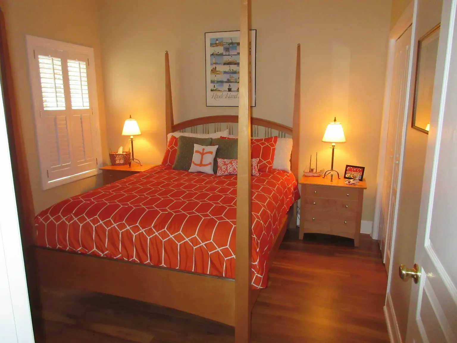 Bedroom with a wooden four-poster bed, orange patterned bedding, nightstands, and a window with shutters.