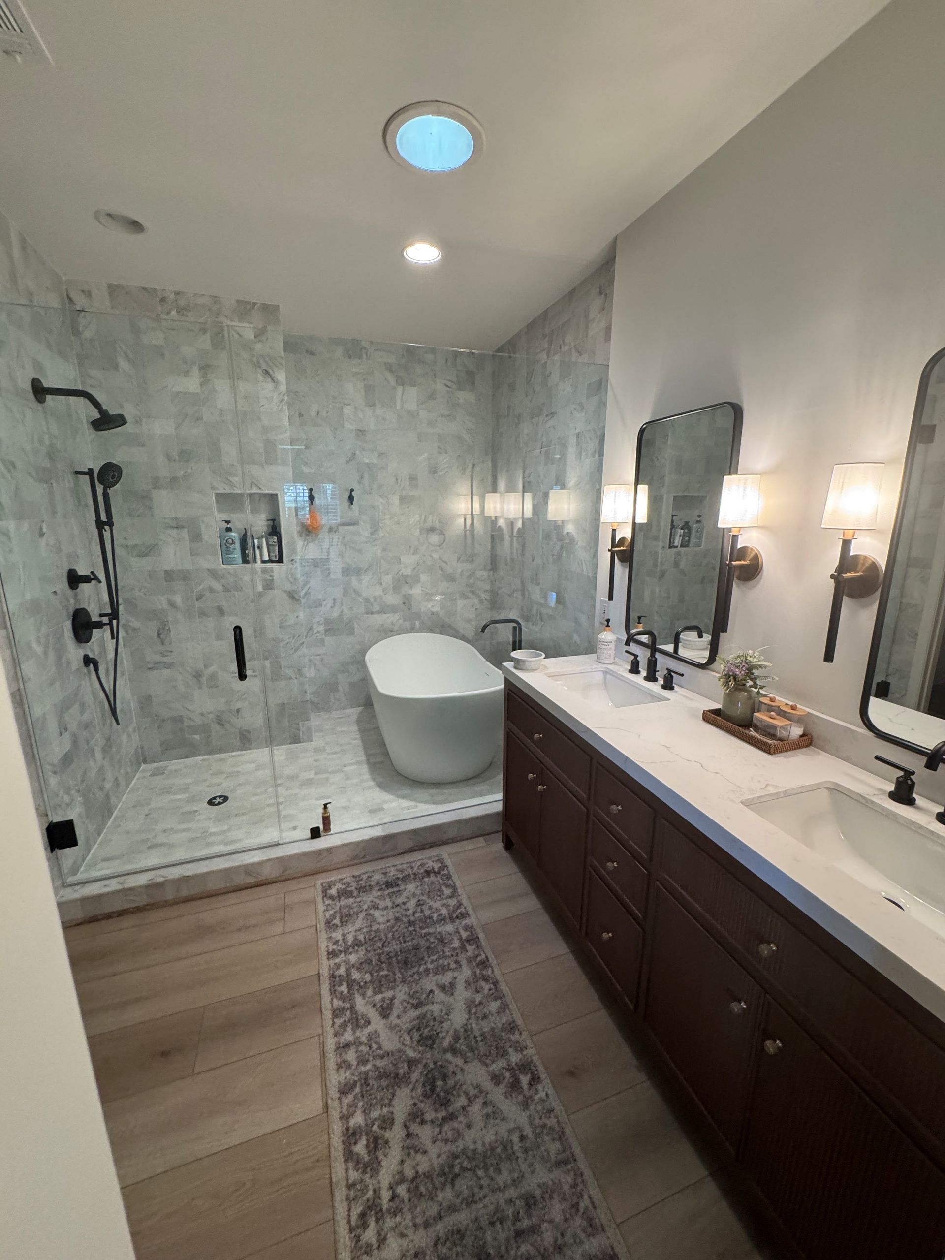 Bathroom with walk-in shower, soaking tub, double vanity, and gray marble tile. Brown cabinets, black fixtures.