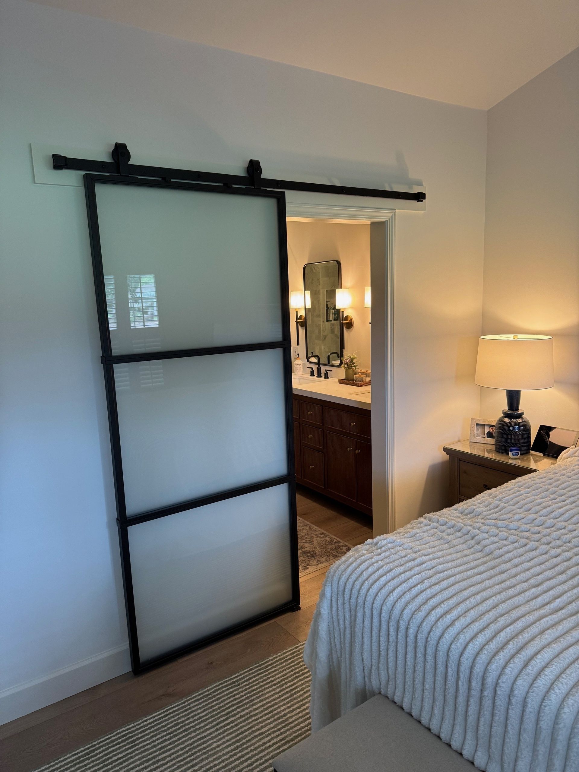 Sliding frosted glass door with black frame, leading to a bathroom. Bedroom with lit lamp and bed.