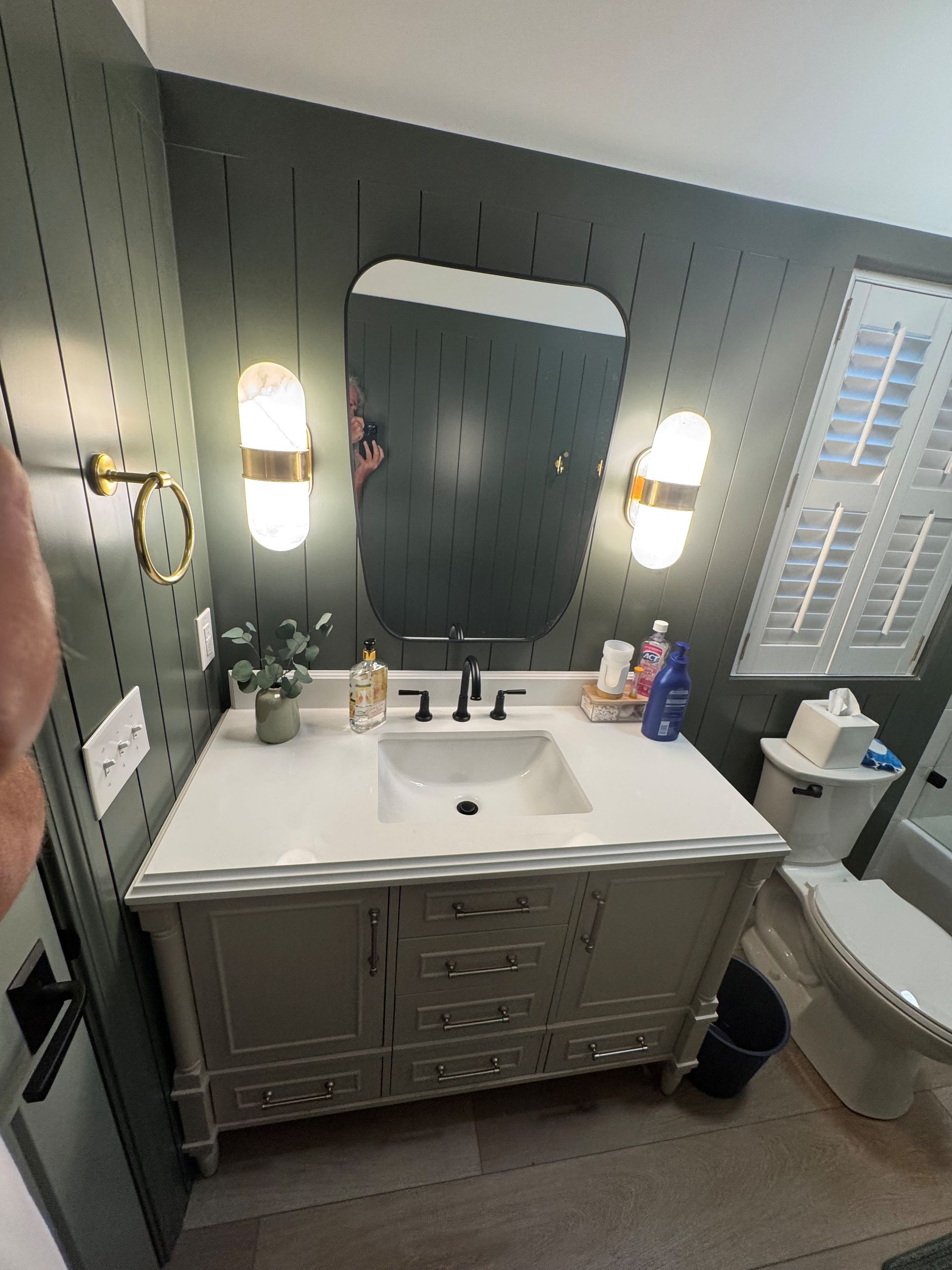 Bathroom with gray vanity, black faucet, oval mirror, and sconce lighting. Gray beadboard wall.