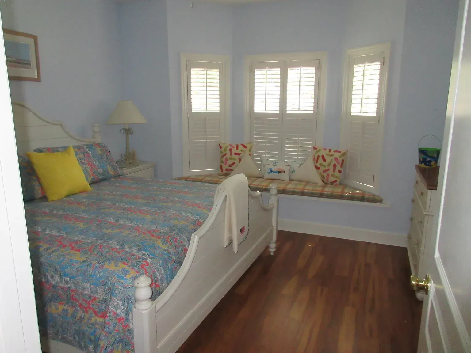 Bedroom with blue walls, white bed, bay window with bench seat, wood floor.
