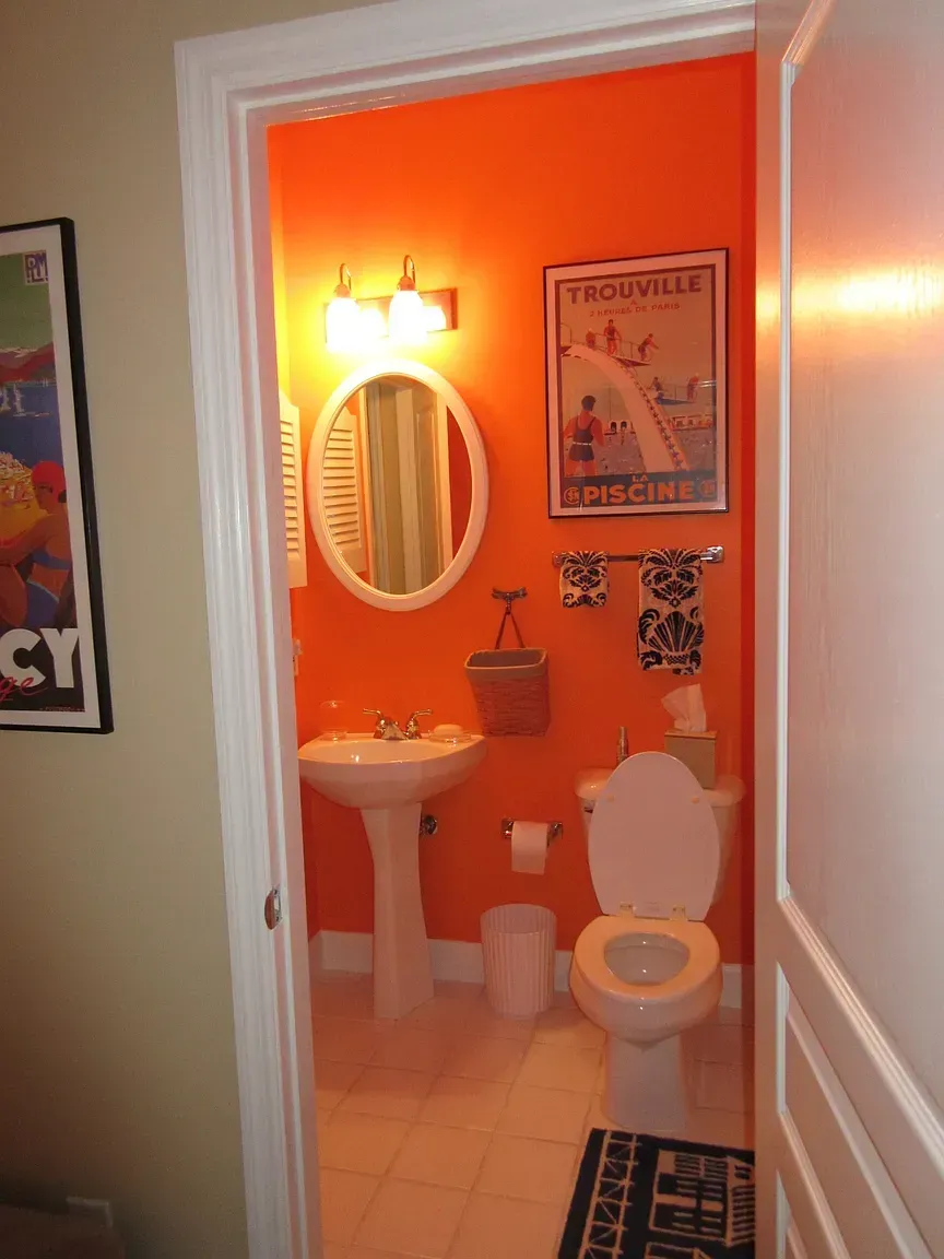 Orange-walled bathroom, viewed through doorway. Includes toilet, sink, mirror, artwork, towel rack, and waste bin.