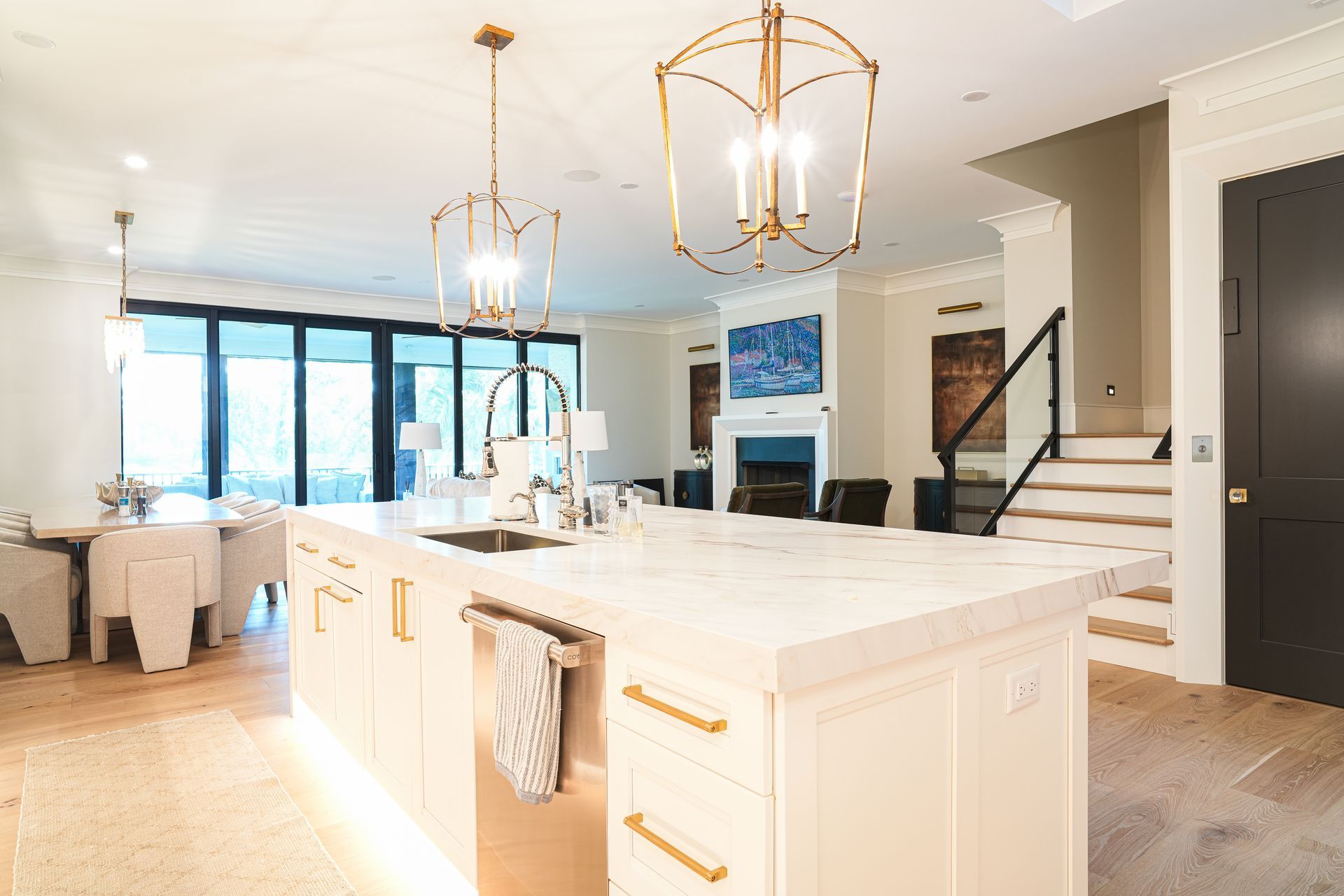 Modern kitchen with white island, gold light fixtures, dining area, and stairs.