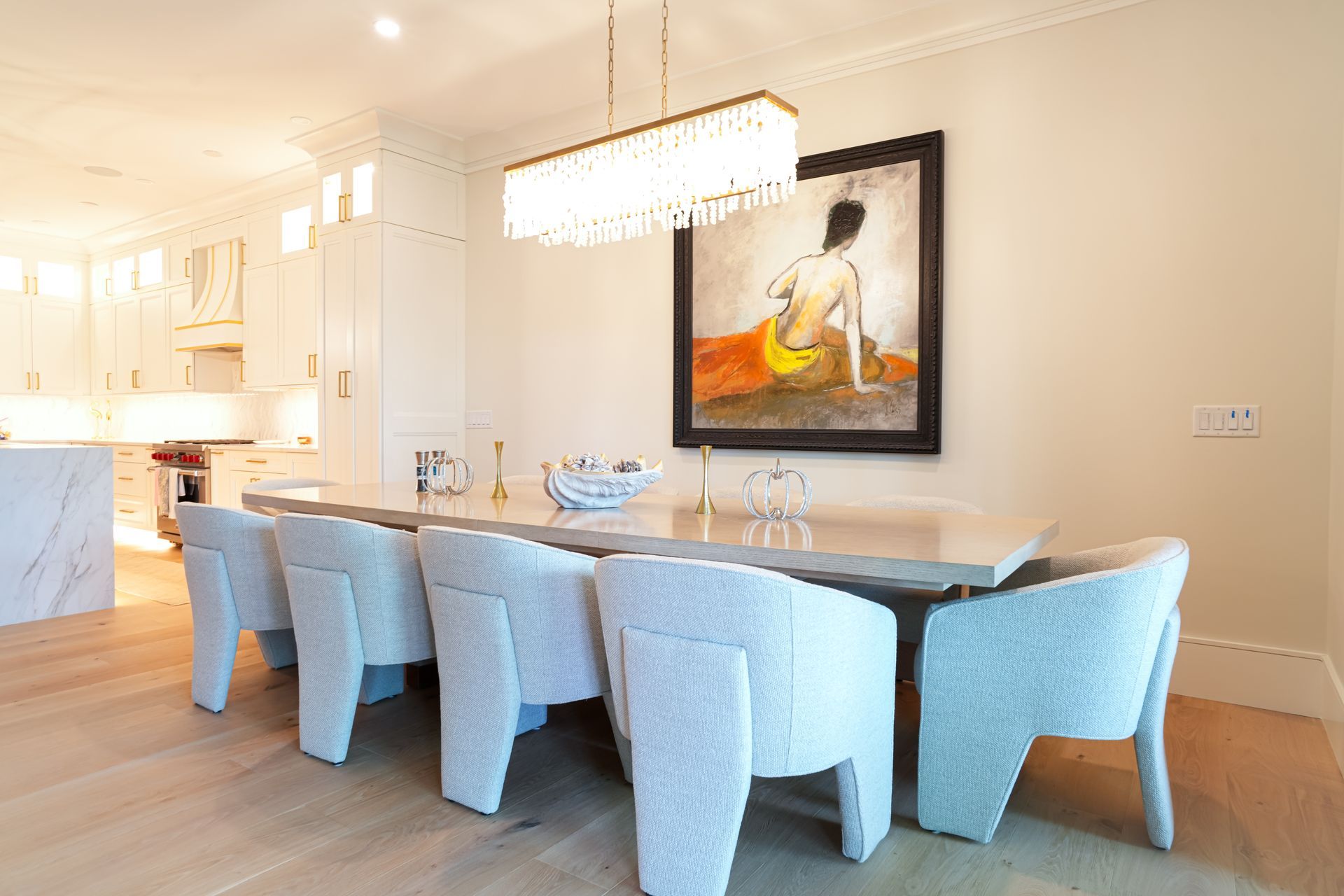 Dining room with white chairs, long table, art, and chandelier.