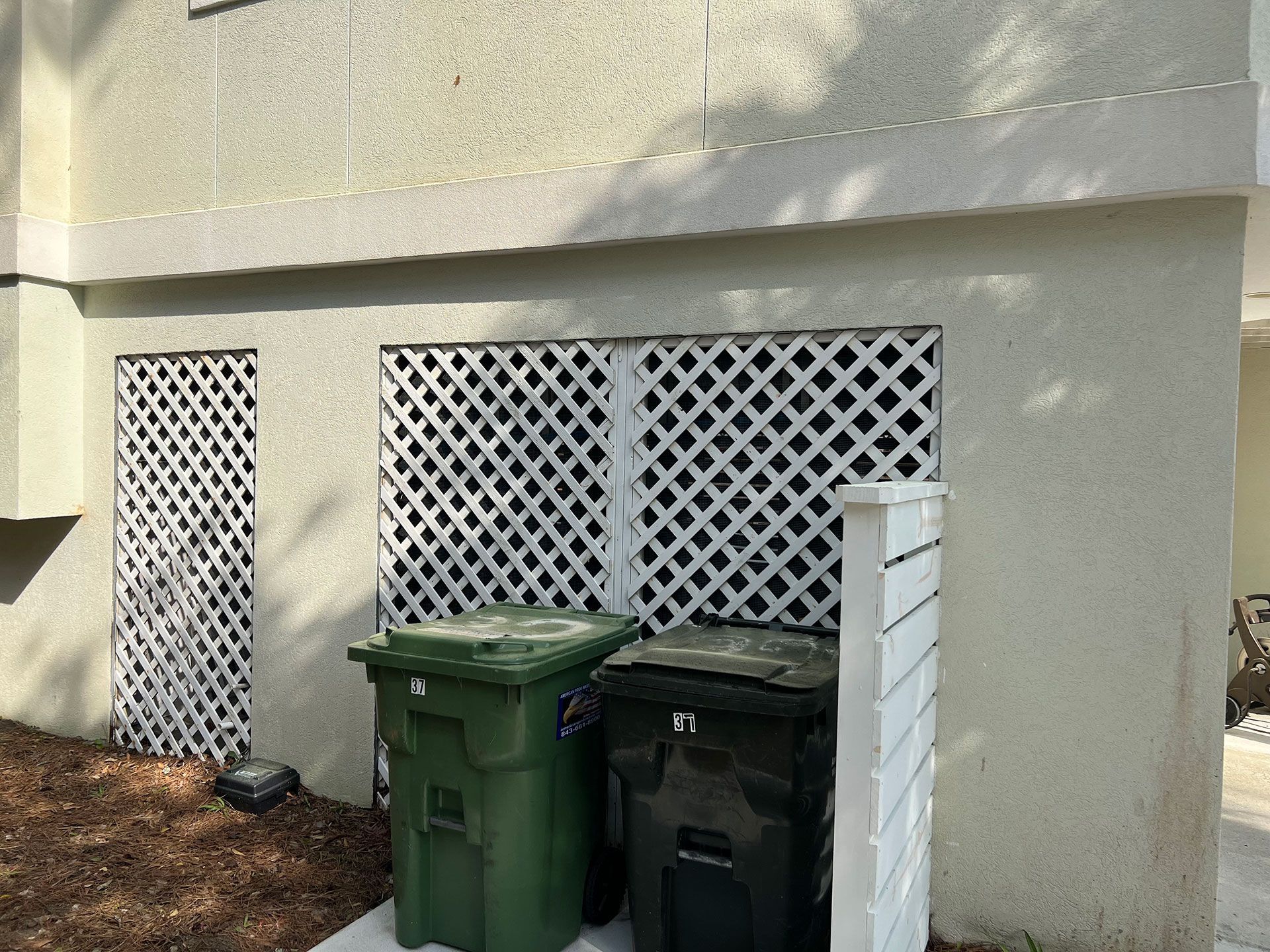 White lattice covers openings in a building's exterior. Two trash bins are below.