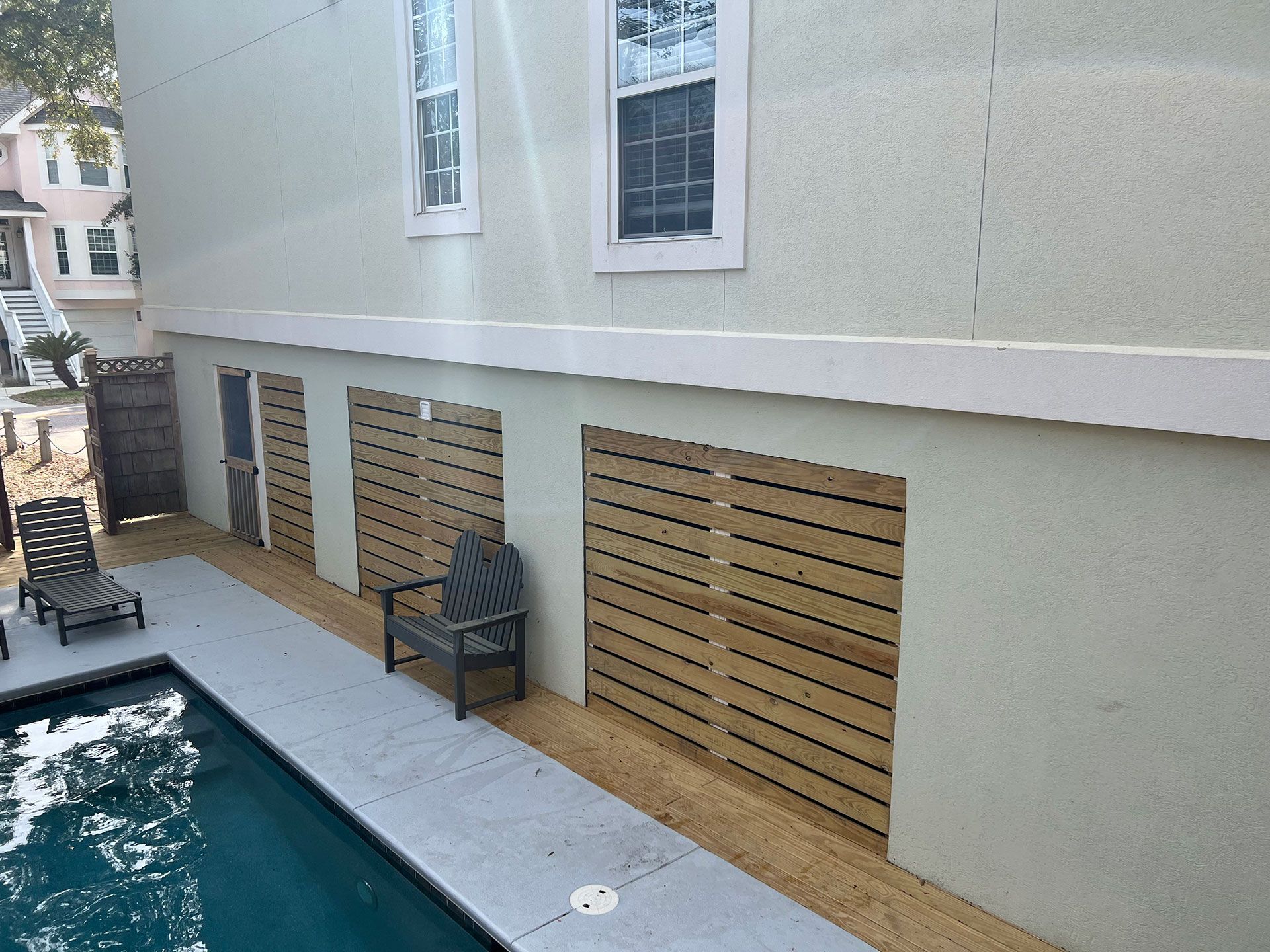 Exterior view of a house with a pool. Wooden slat panels cover openings in the wall, with seating and a walkway.