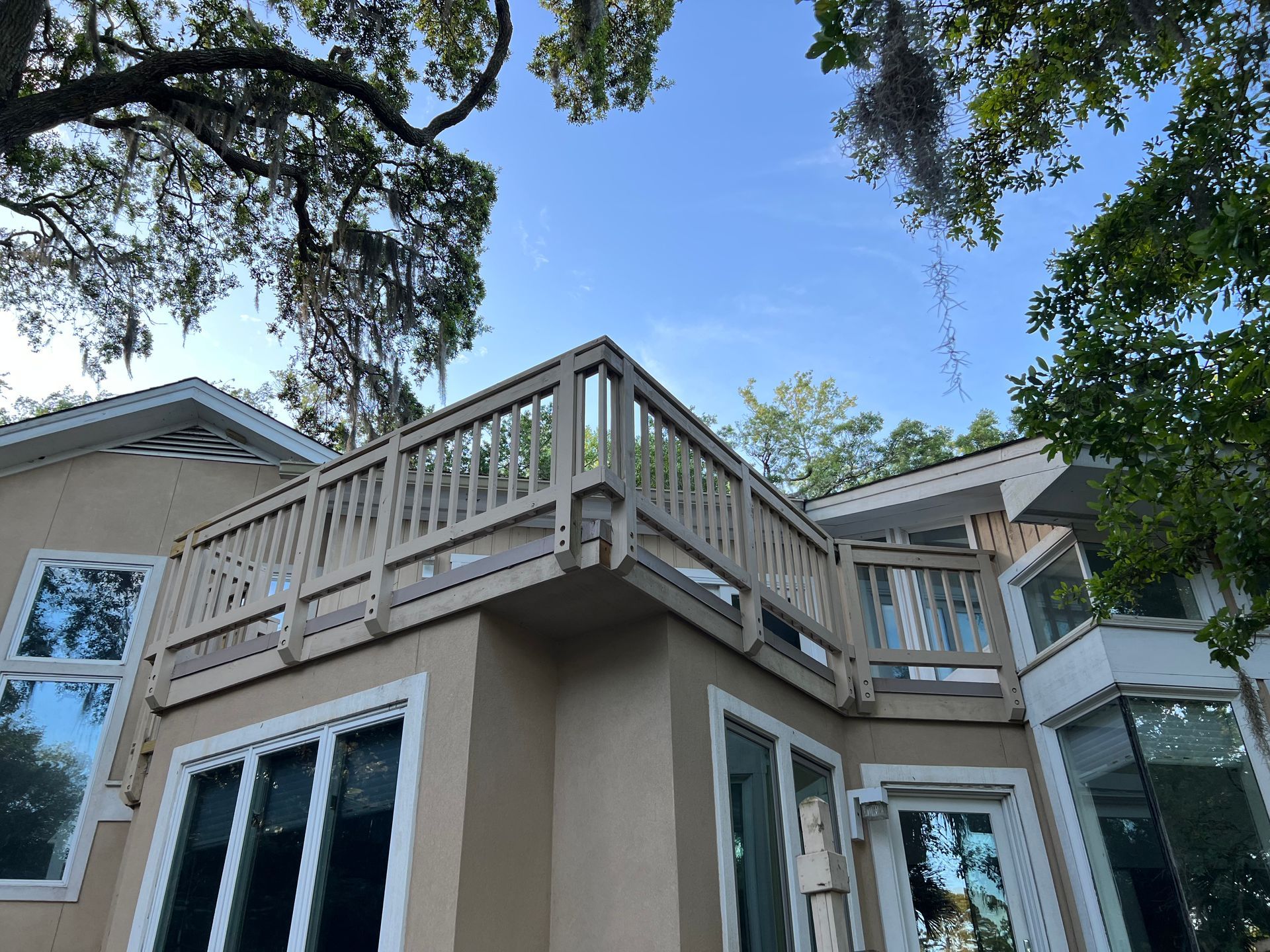 Tan house with a wooden deck and railing. Blue sky peeks through trees overhead.