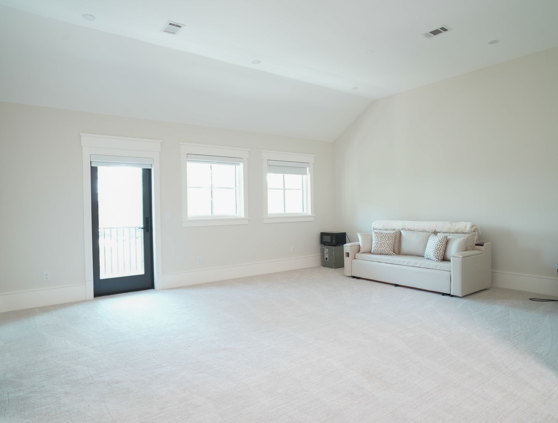 Empty room with white walls, carpet, and windows; a sofa sits against one wall.