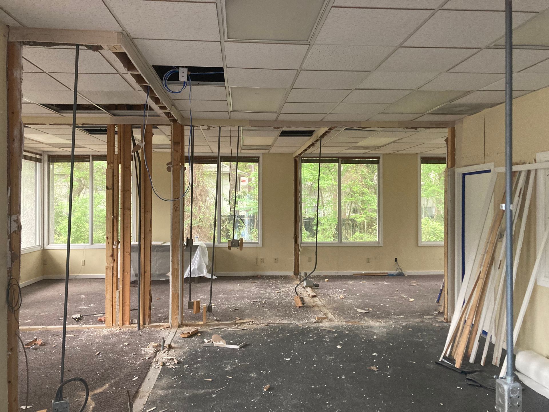 Interior demolition of a room; exposed framing, ceiling tiles removed, windows, and debris on the floor.