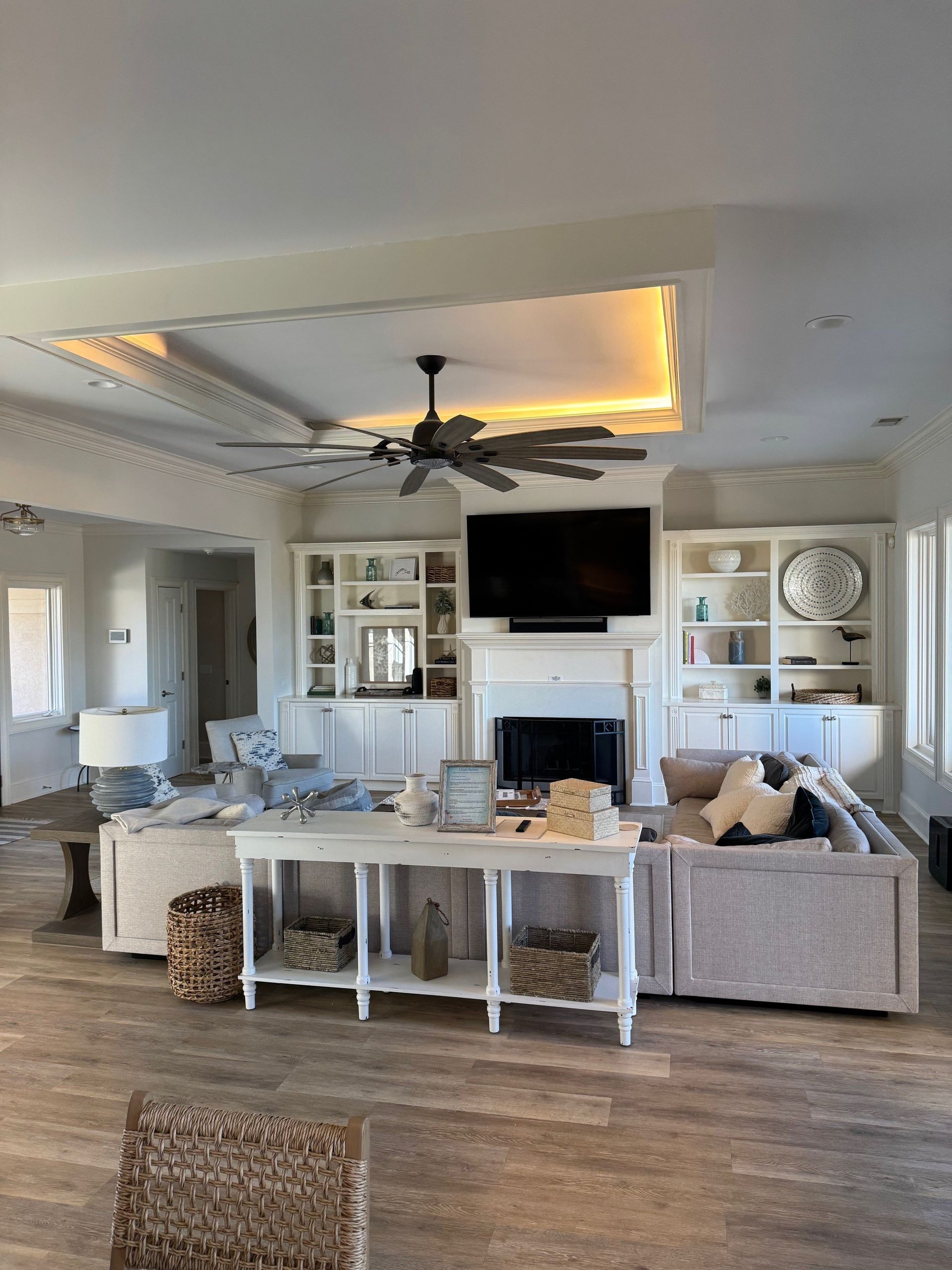 Living room with white built-ins, fireplace, large TV, and ceiling fan. Gray and white furniture, wood floors.