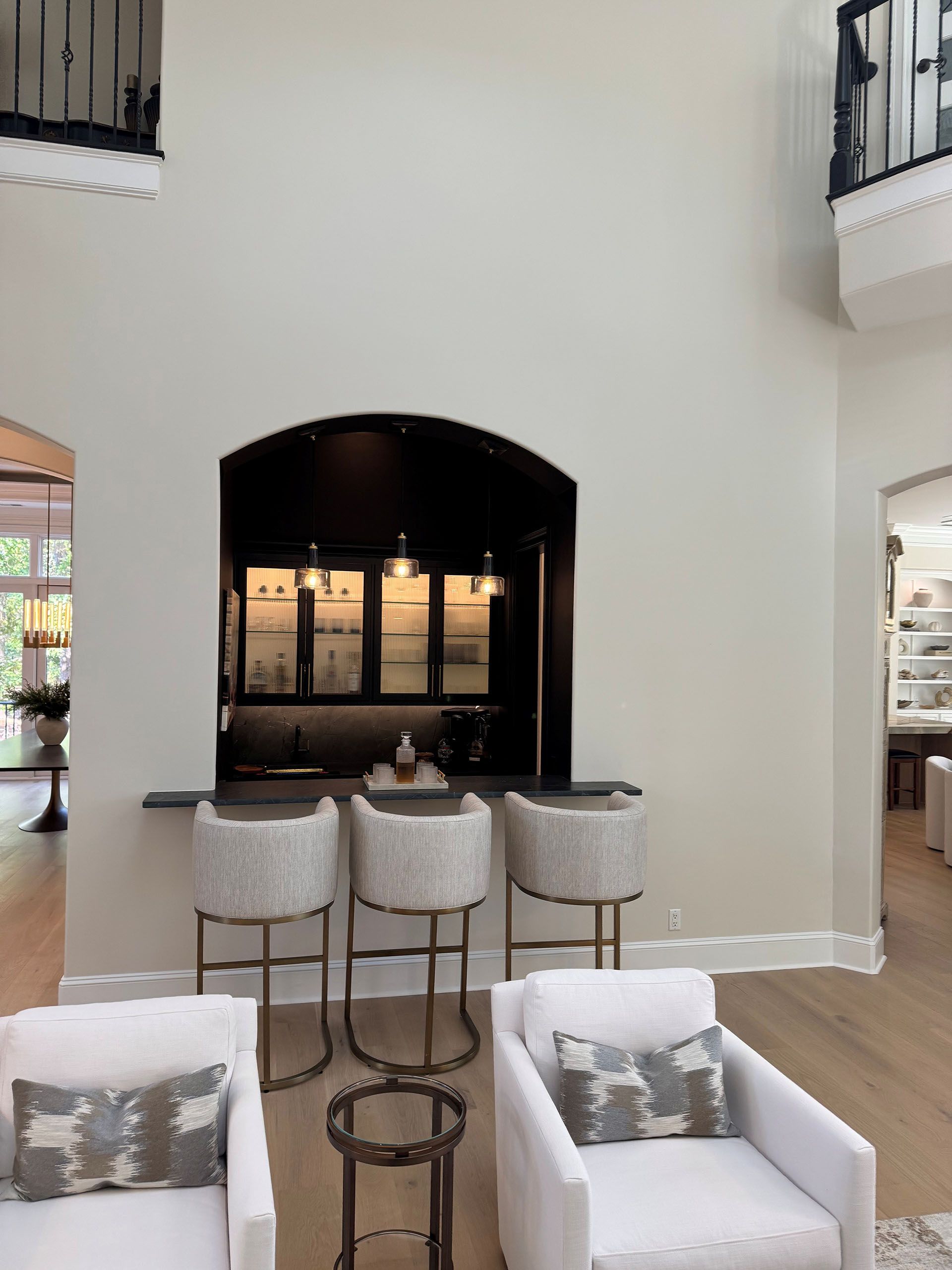 Wexford Home Remodel - After: Interior with a bar, stools, and white armchairs. High ceilings, neutral color palette, and natural light.