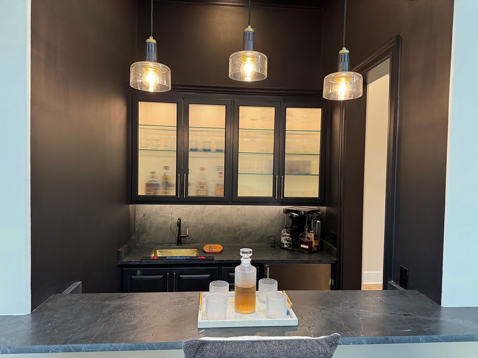 Wexford Home Remodel - After: Dark home bar with black cabinetry, countertop, and pendant lights. A liquor decanter and glasses are on a tray.