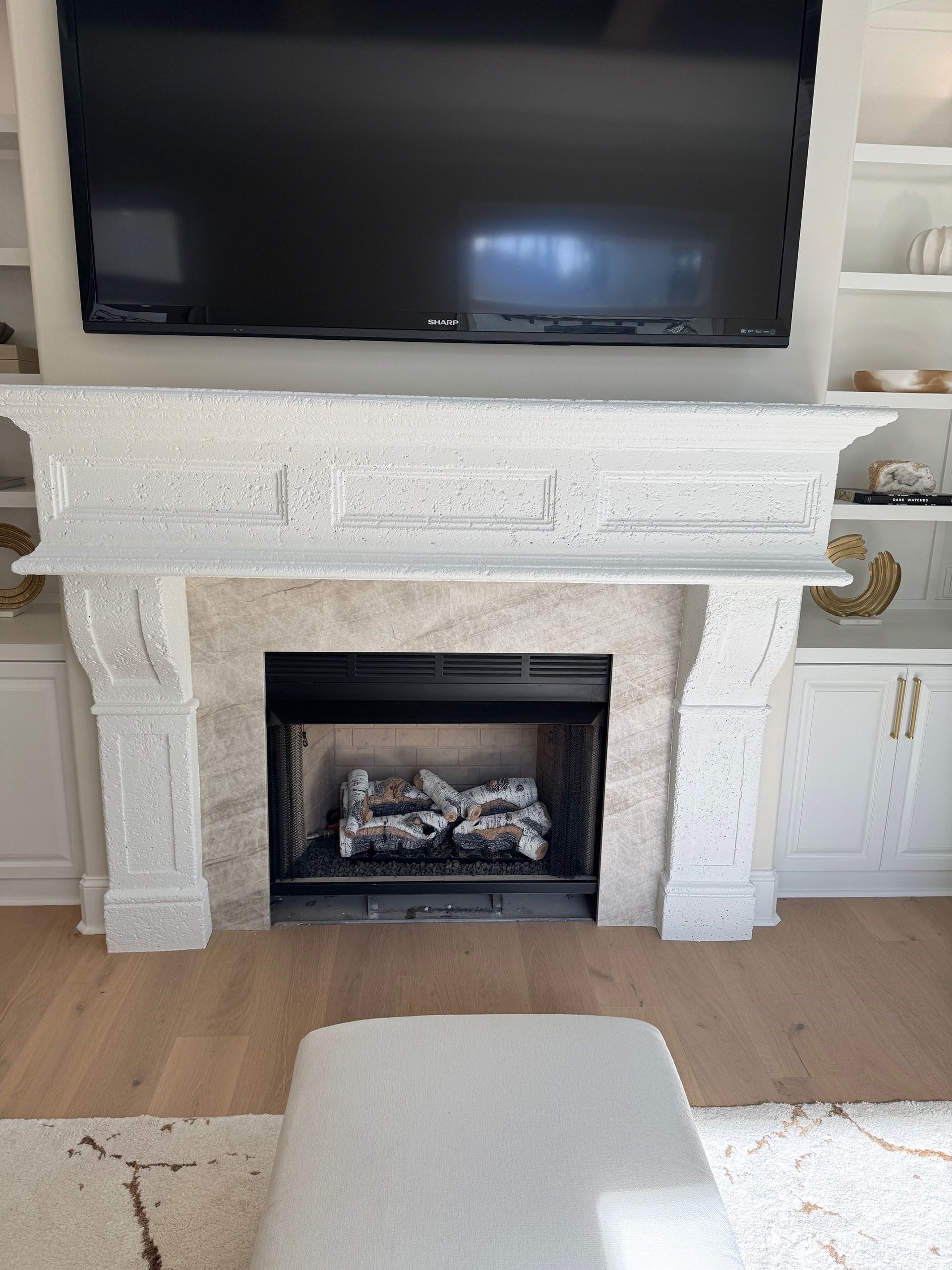Wexford Home Remodel - After: Fireplace with TV above, flanked by white built-in shelves. A white ottoman sits in front.