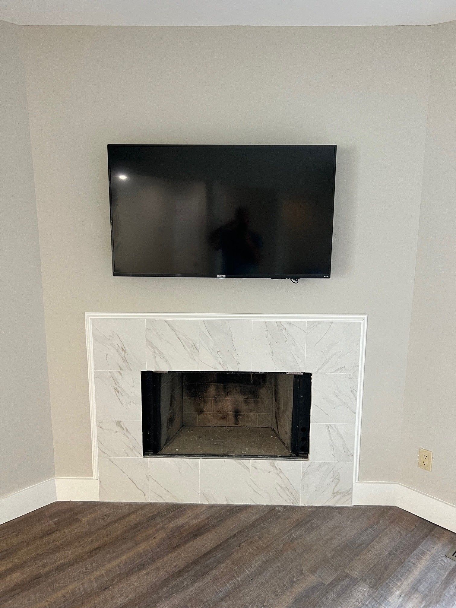 TV mounted above a marble fireplace with a dark interior. Room with light walls and dark wood floor.