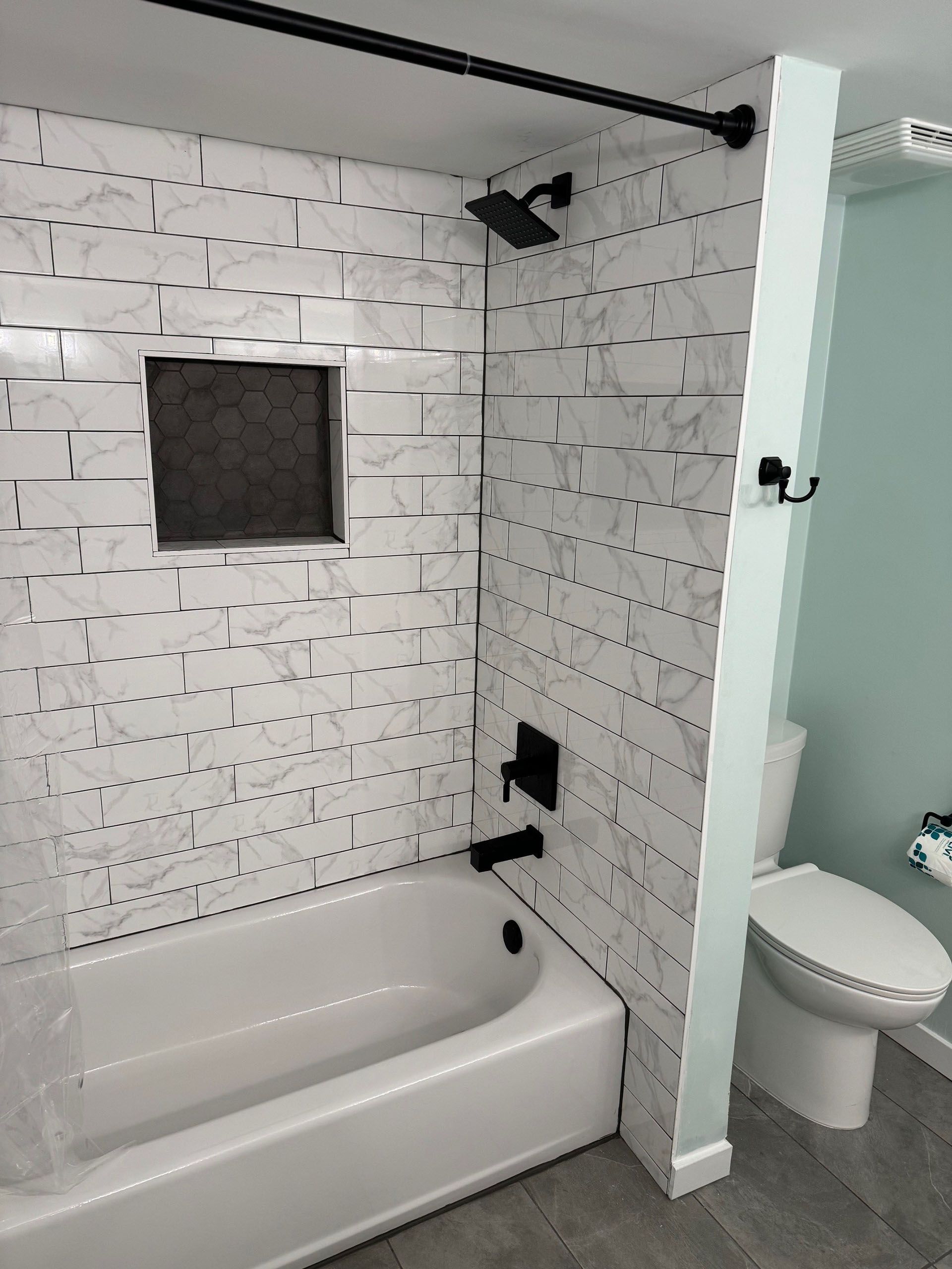 Miscellaneous Bathroom Projects - Bathroom with white marble-like tile, a black showerhead, and a white tub next to a toilet.