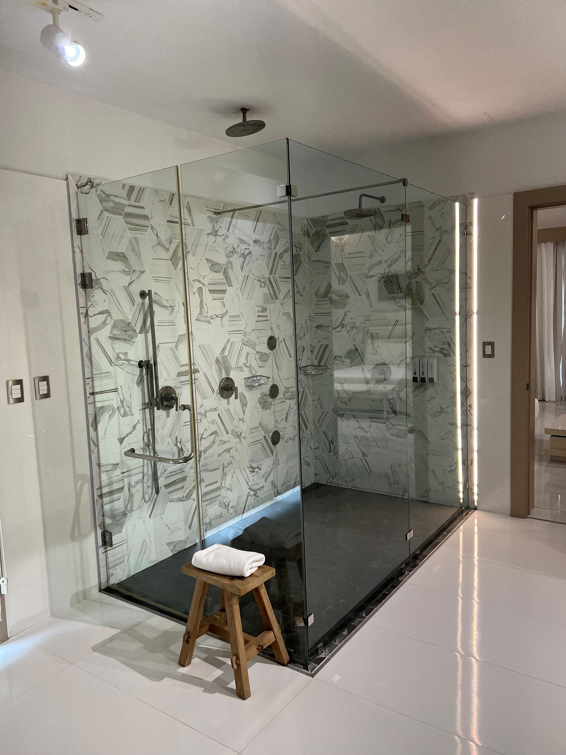 Miscellaneous Bathroom Projects - Glass shower enclosure with patterned wall, dark floor, and wooden stool in a white-tiled bathroom.