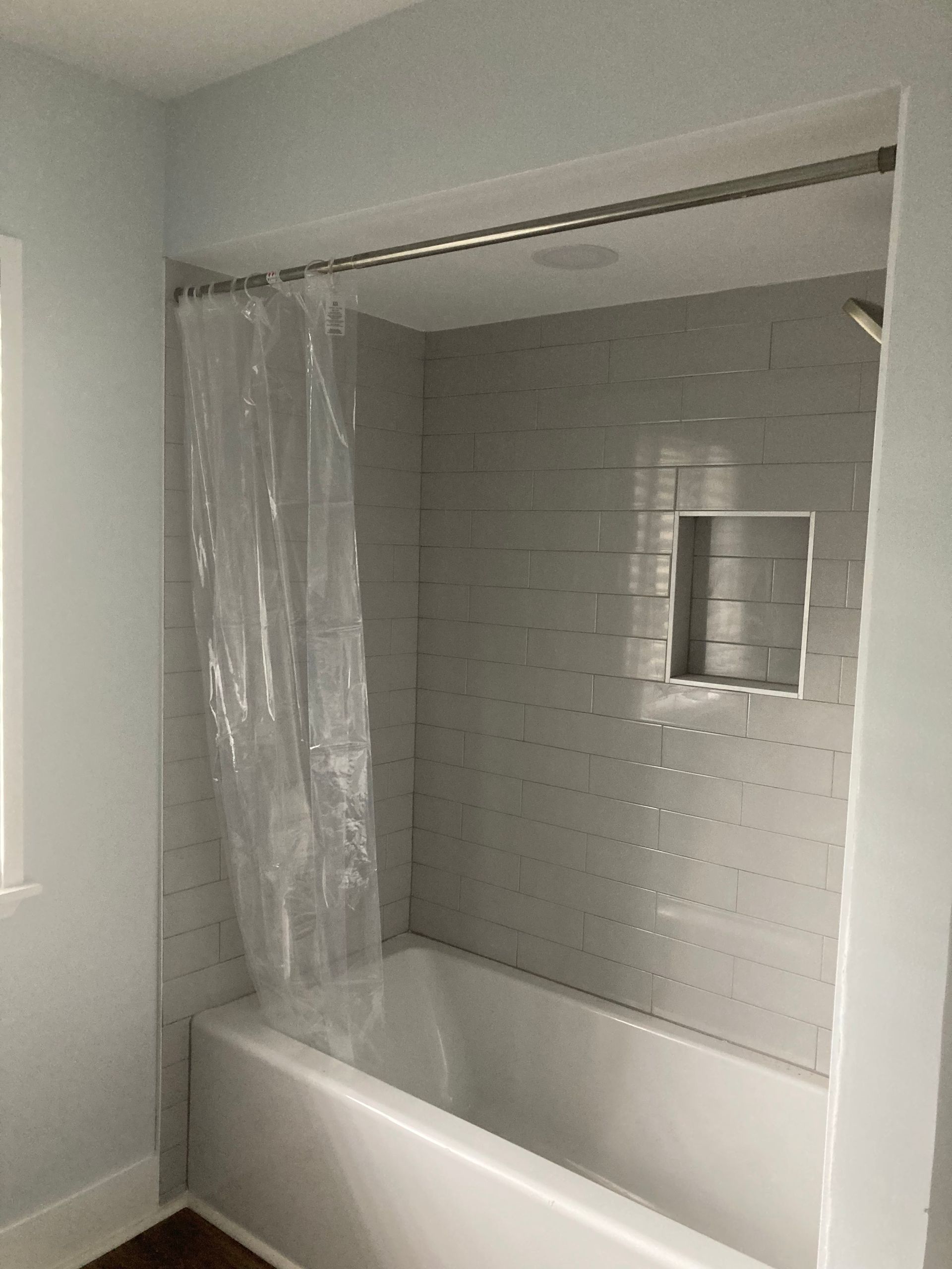 Bathroom with white tub, gray tiled walls, clear shower curtain, and a recessed niche.