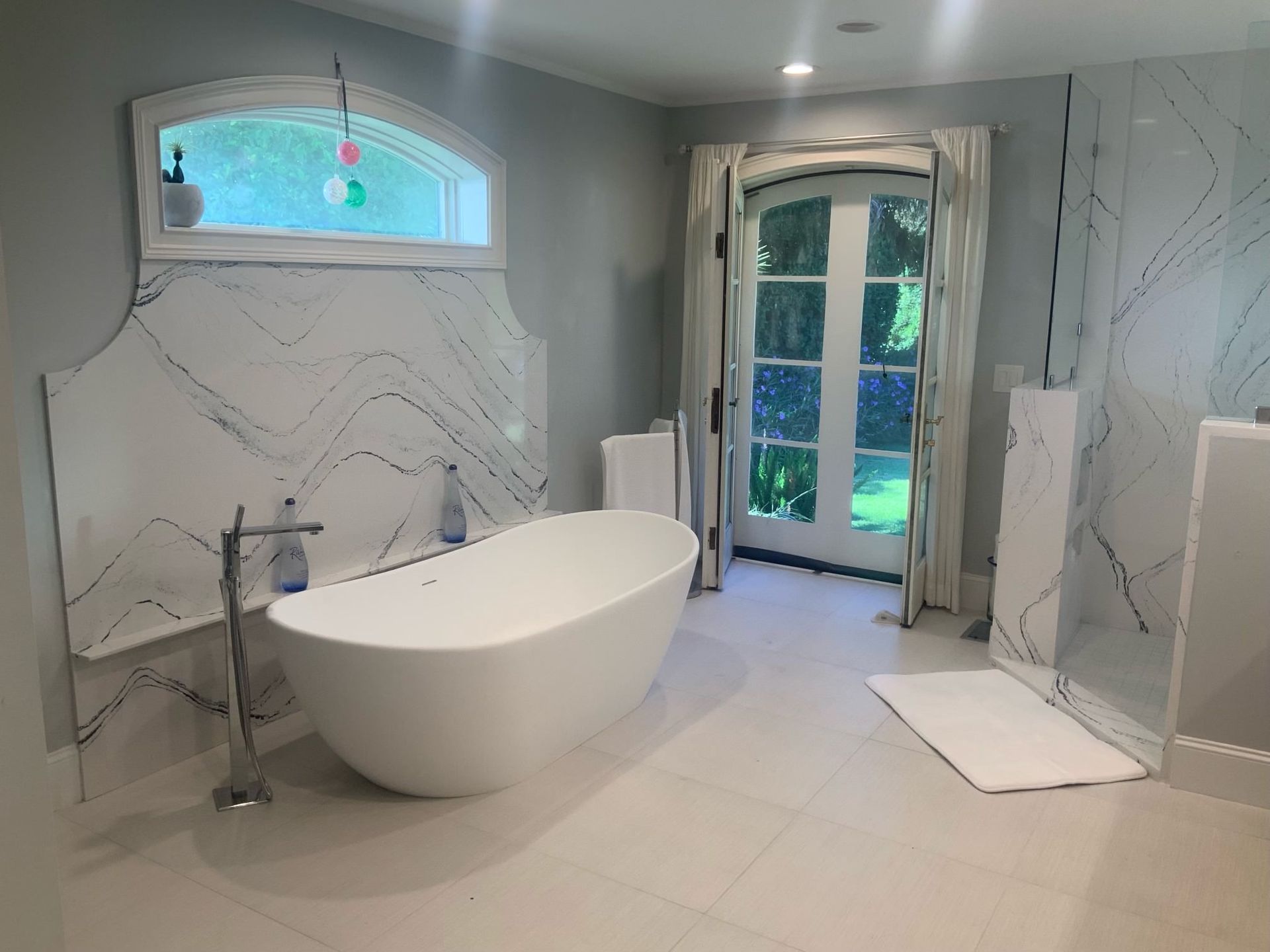 Spacious white bathroom with freestanding tub, marble accent wall, and French doors to outdoors.