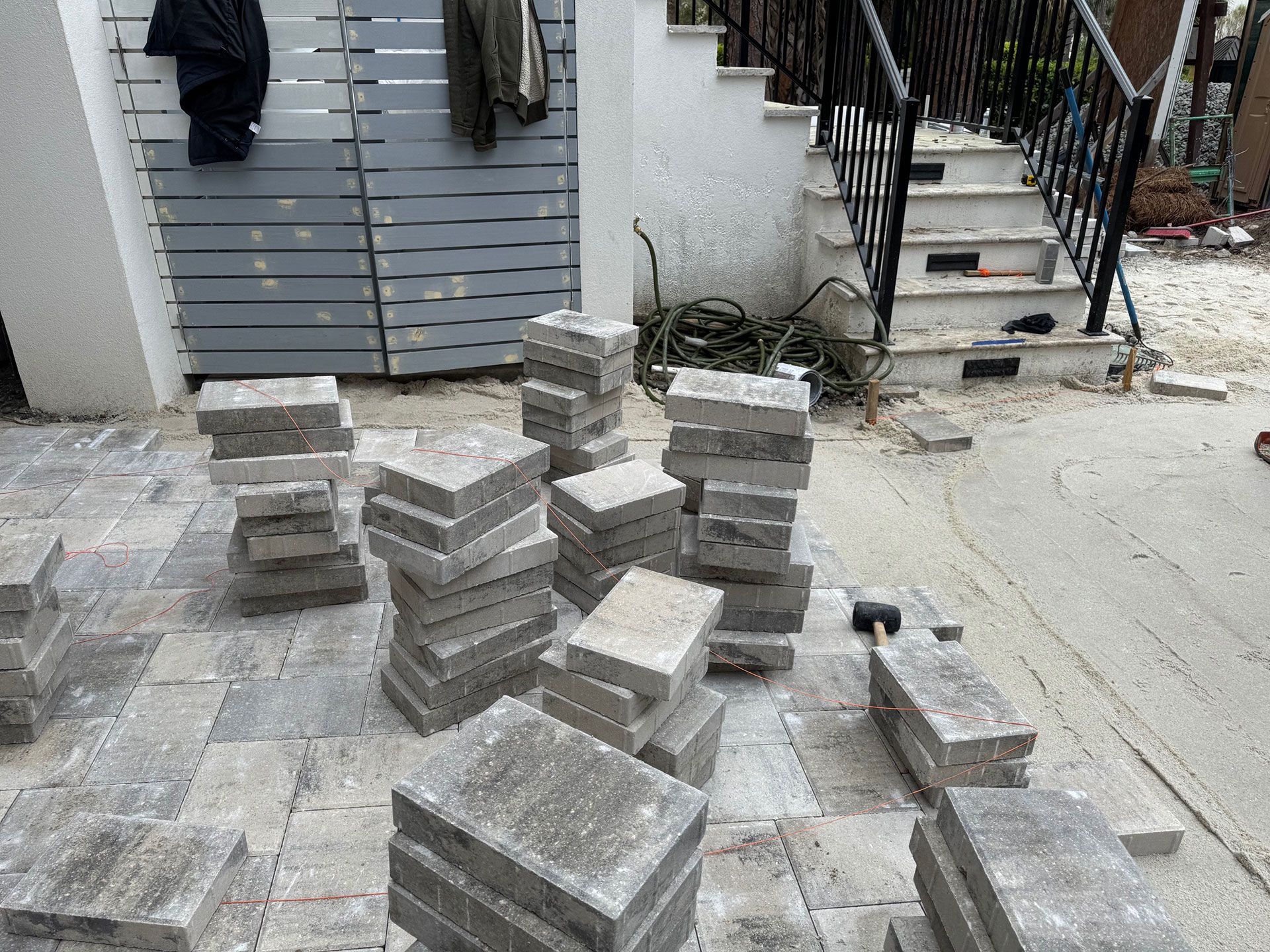 Piles of gray paving stones on patterned pavement; stairs and a wall are in the background.