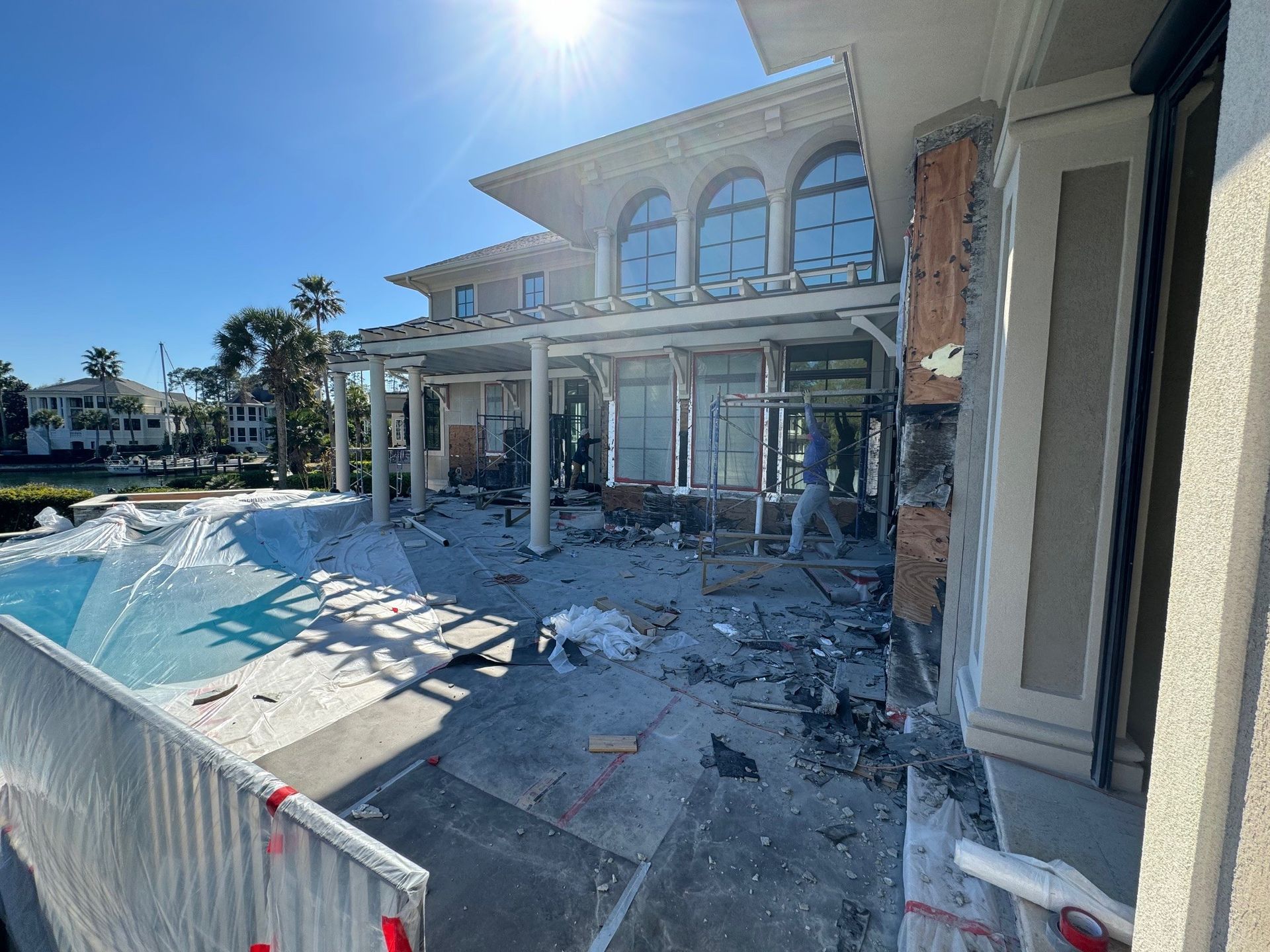Exterior view of a house with a pool undergoing construction, debris visible. Blue sky and sunlight.