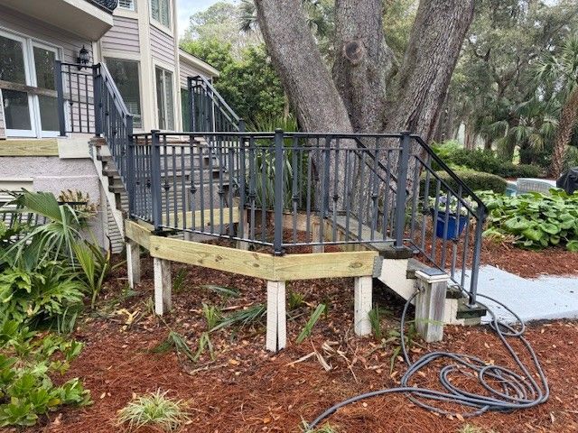 Wooden deck with black metal railing and steps, built on stilts, surrounded by mulch and landscaping.