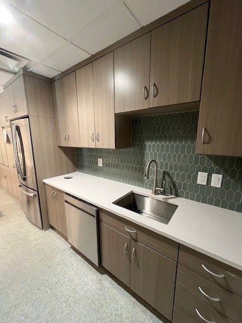 Modern kitchen with light brown cabinets, white countertop, stainless steel sink and appliances. Teal tile backsplash.