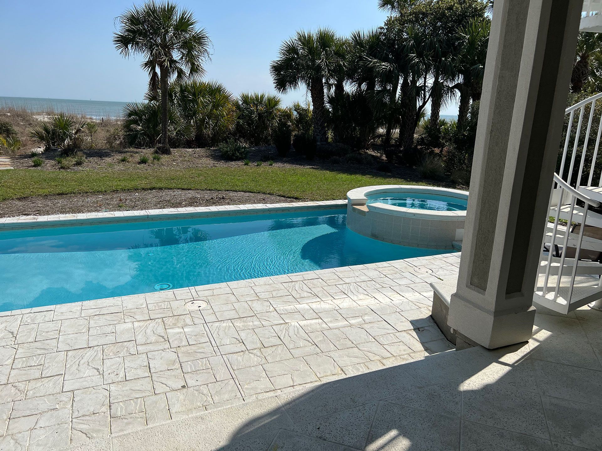 Swimming pool and hot tub overlooking a beach; blue water, white patio, and palm trees.