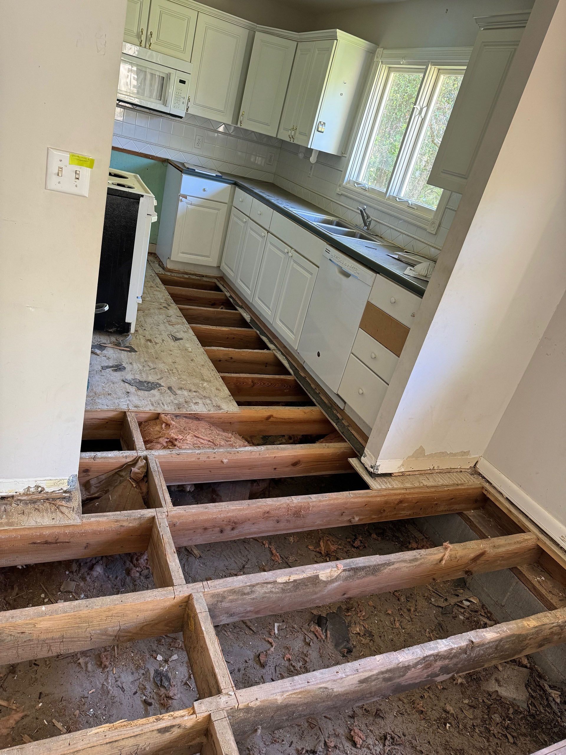 Termite Damage - Interior view of a kitchen under renovation, with exposed floor joists. White cabinets, black countertop, and a window are visible.