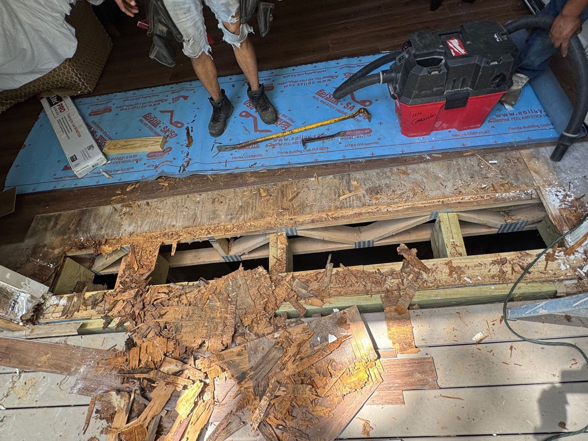Water Damage - Damaged wall with exposed wood framing. Two people and tools near blue surface.