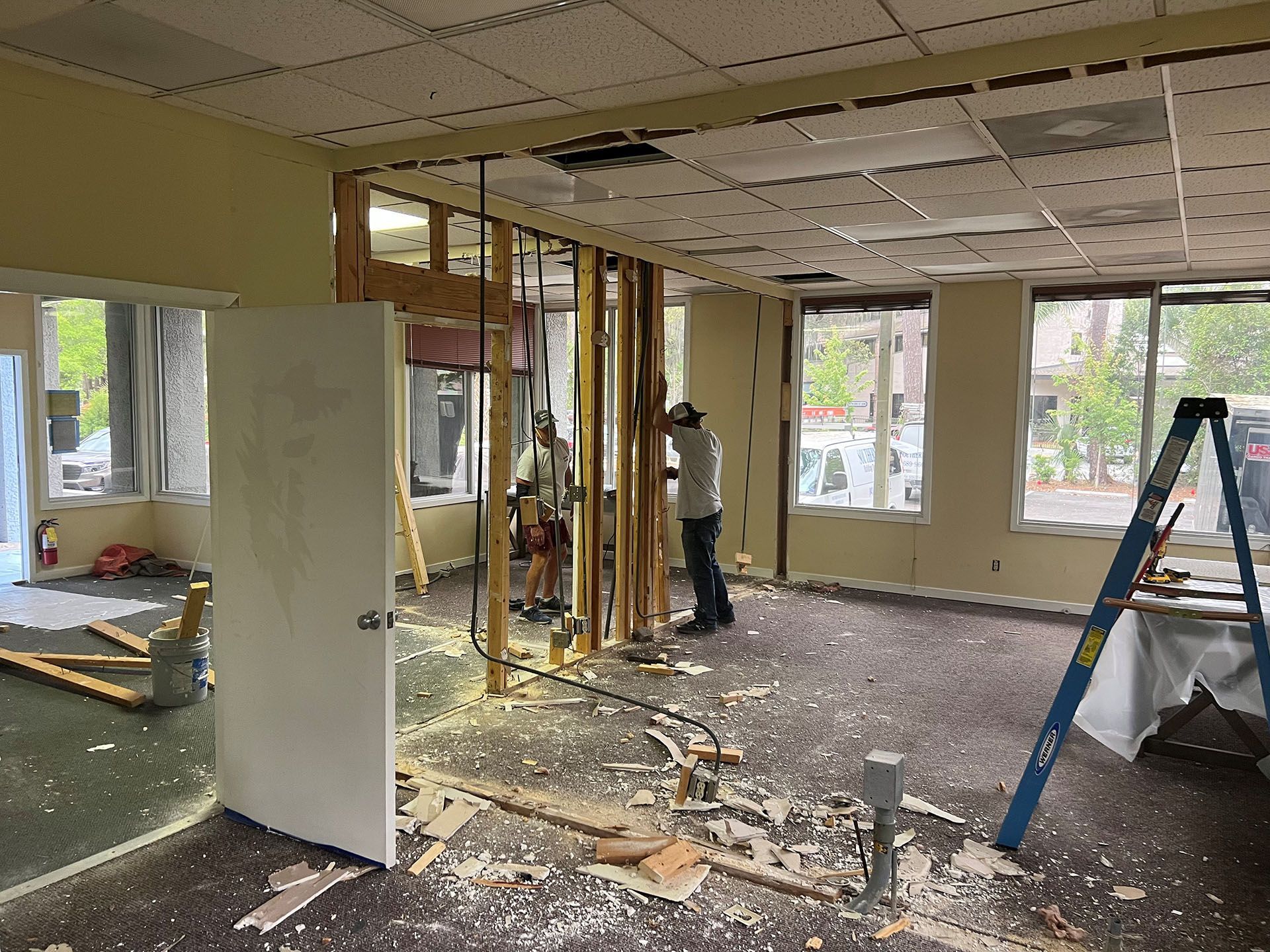 Carolina Rides - During: Interior during renovation; workers dismantling a wall in a room with windows, debris on the floor, and a ladder.